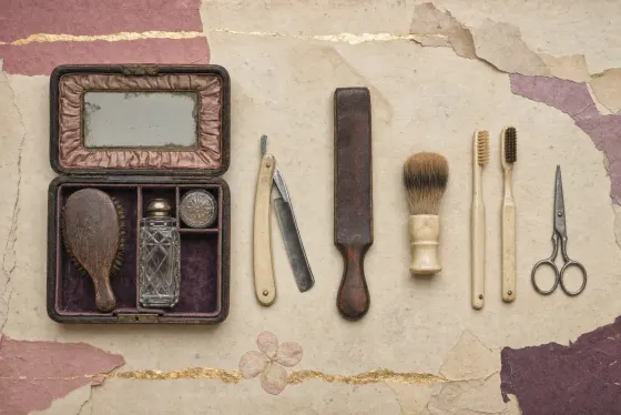 A Victorian man's dressing case of dark leather lying open, its lid lined with gathered pink silk around a mirror and its compartments holding a wooden hairbrush, a cut-glass scent bottle and a small round pot, with a folding razor, a leather strop, a shaving brush, two toothbrushes and small scissors laid out in a row beside it.
